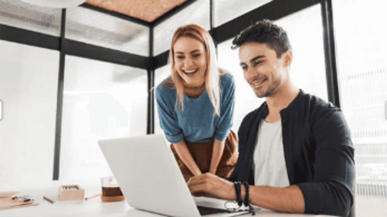 Two coworkers hover over a laptop with smiles on their faces
