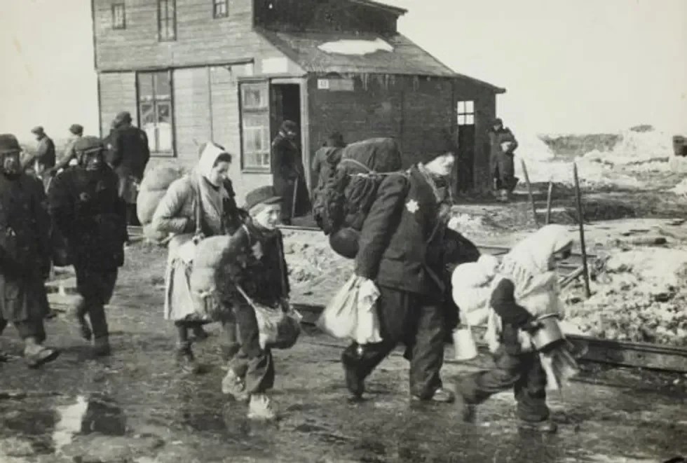 Boy walking to deportation in a group, wearing cap, satchel and backpack.