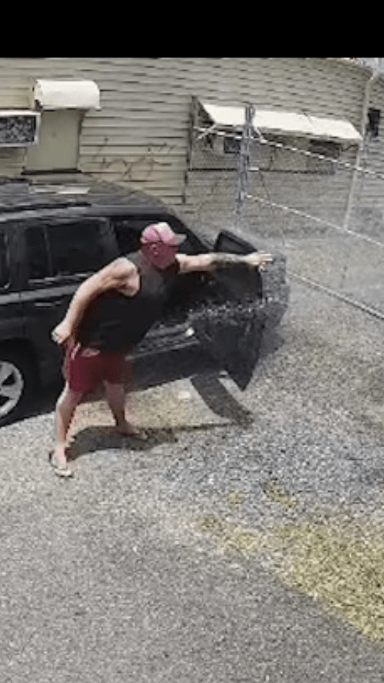 man in black tank top and red shorts stands beside his car with the door open trying to block a spray of water
