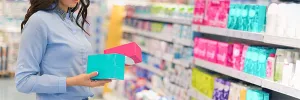 A woman stands in a drugstore aisle, choosing between two brands of tampons. Public health, Regulation, Menstruation, Personal care products, Heavy metals, Tampons, Toxic shock syndrome, PFAS, Phthalates, Toxic chemicals, Period products, Menstrual products, Women's health care