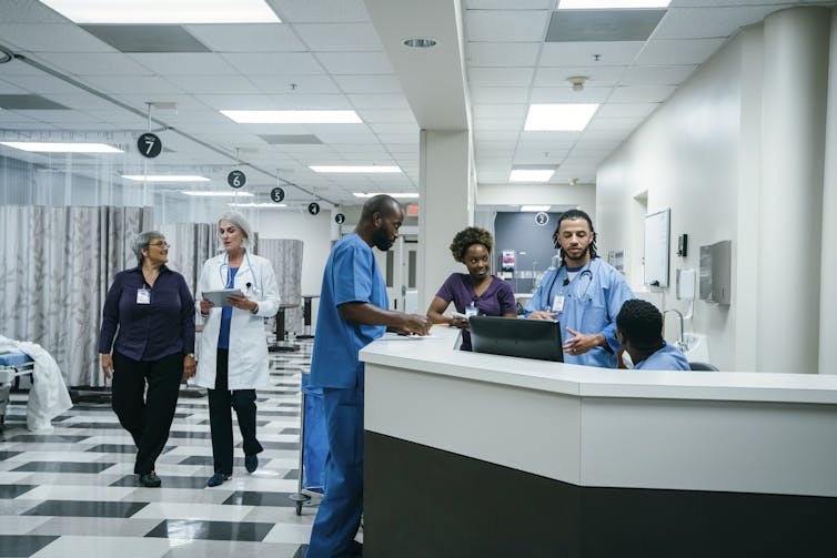 Black doctor in blue scrubs talks with medical team at nurse's station.
