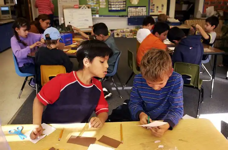 Two boys sit next to each other at a desk in a classroom filled with other children at desks.
