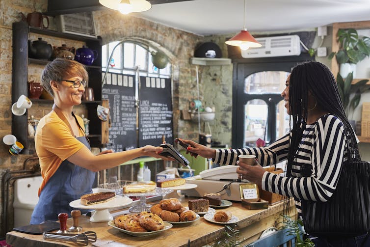 A young woman with dreadlocks pays for her coffee as a smiling young female barista with short hair holds out a card reader.