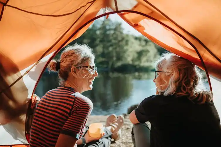 Two women with gray hair and glasses sit inside a tent, looking out at a pond, as they smile and chat.