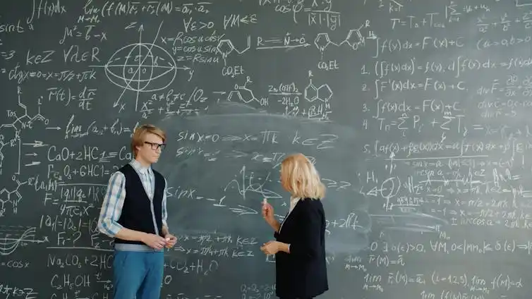 A teacher talks to a student before a chalkboard wall filled with equations, chemical structures and graphs