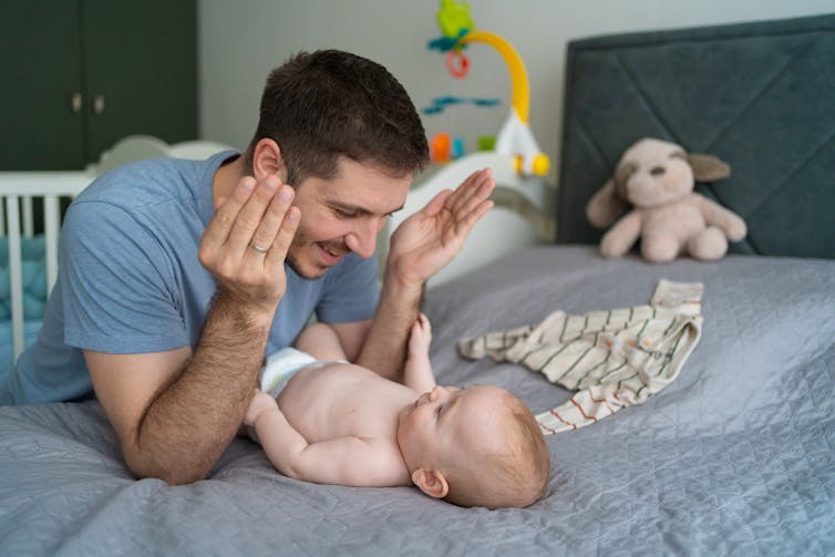 A man holds his hands away from his face and leans over a small baby lying on a bed and smiles.