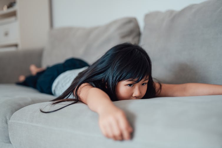 A young girl with dark hair lays on her stomach on a couch with her arms and legs splayed out.