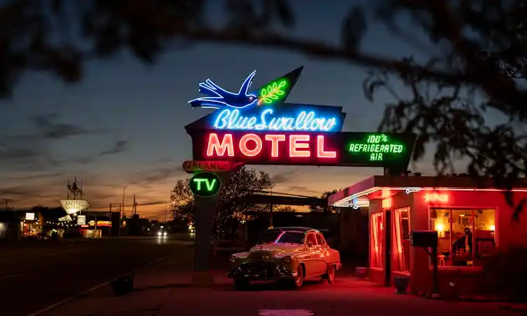 A roadside motel at dusk with a vintage car parked in the lot and a large blue and pink neon sign reading 'Blue Swallow Motel.'