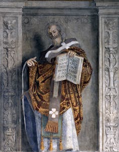 A bearded figure in an orange and brown robe gestures toward a large open book that he holds in one hand.