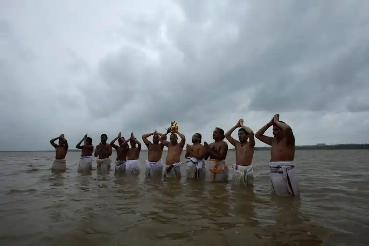 A line of around a dozen men wearing white sarongs stand in waist-high water, holding their hands up in prayer.