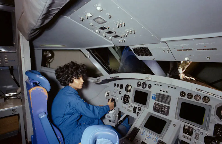 A woman wearing a blue jumpsuit sitting in the pilot's chair in a space shuttle cockpit simulator