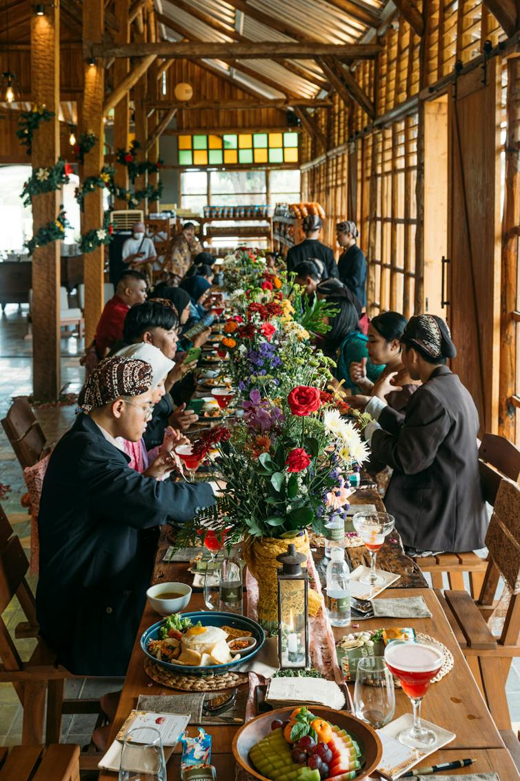 At a long table covered by flowers and plates of food, a diverse group of travelers shares a meal.