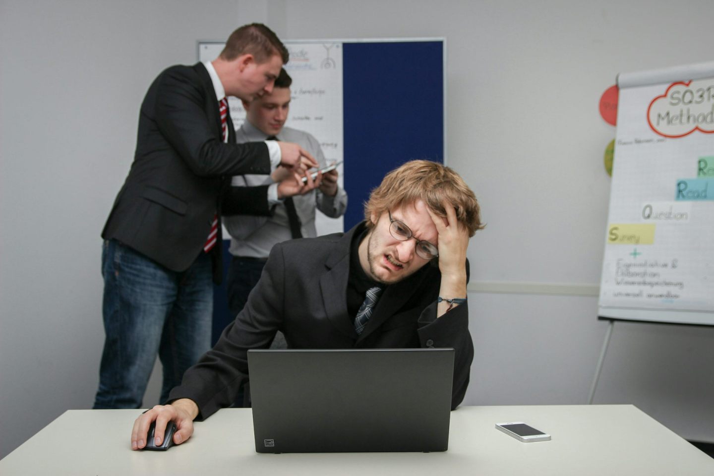 A young man with glasses looks confused and exasperated as he sits in front of a computer, while two colleagues discuss something behind him.
