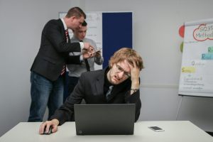 A young man with glasses looks confused and exasperated as he sits in front of a computer, while two colleagues discuss something behind him.