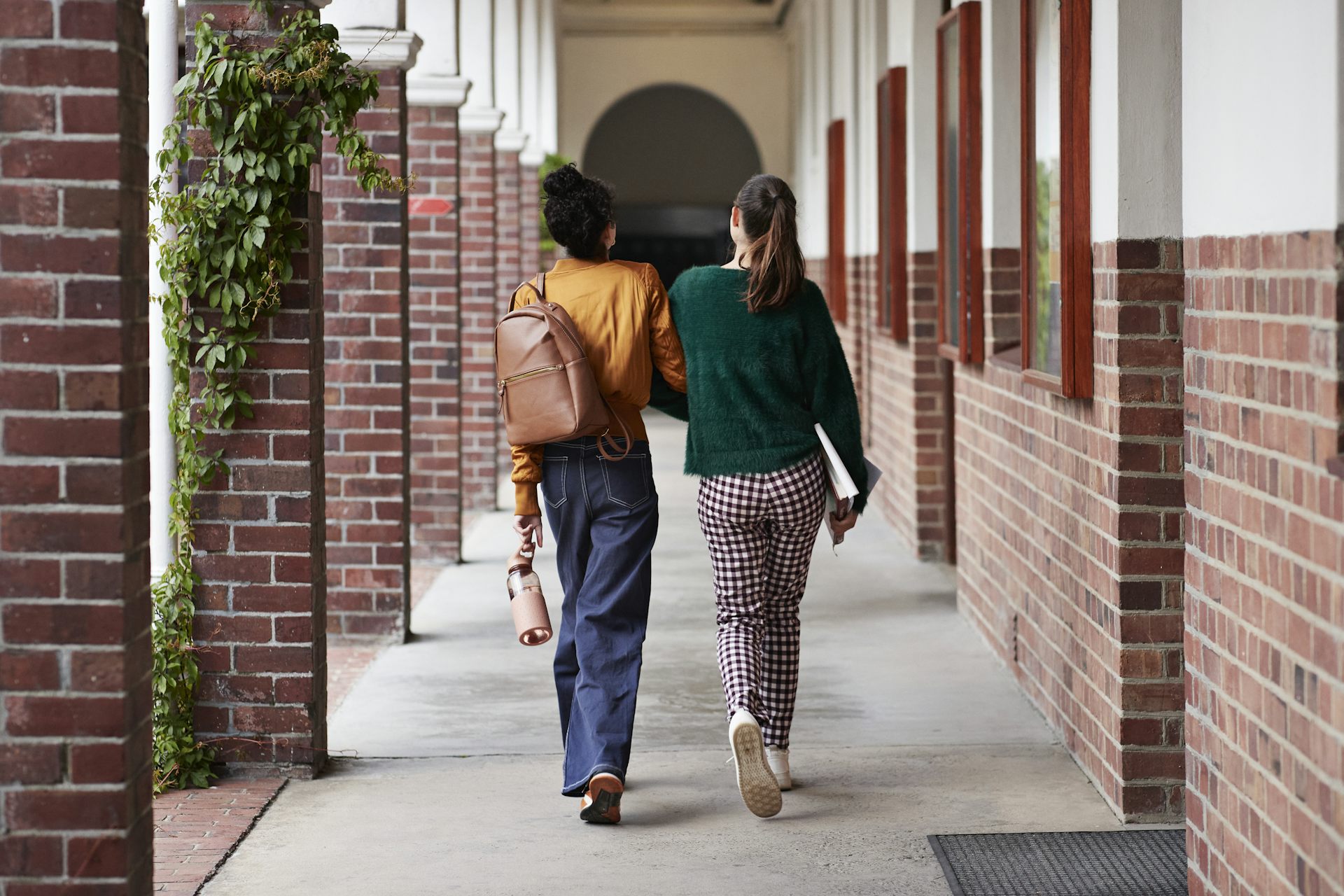 Two women walk closely together down a stone path with red brick pillars on either side of them.