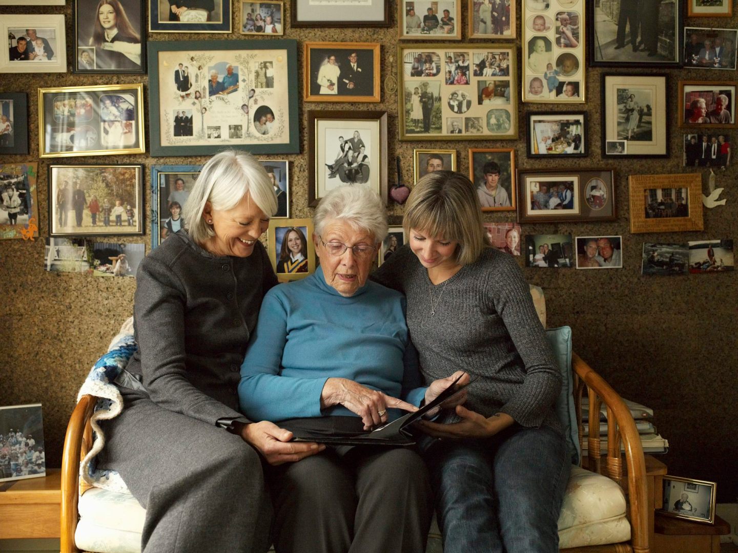 Three generations of women look at a photo album together while behind them a wall features dozens of framed photos.